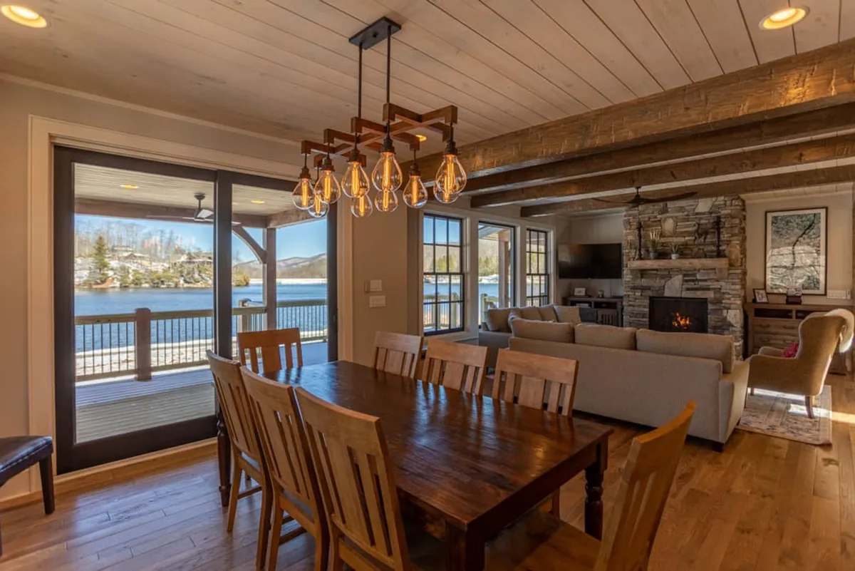 A welcoming dining area is the focus here, with a substantial dining table set beneath a decorative chandelier. Expansive windows overlook the lake, and a fireplace adds warmth to the scene. Comfortable seating nearby invites relaxation, creating an ideal setting for both lively group dinners and quiet, contemplative moments.
