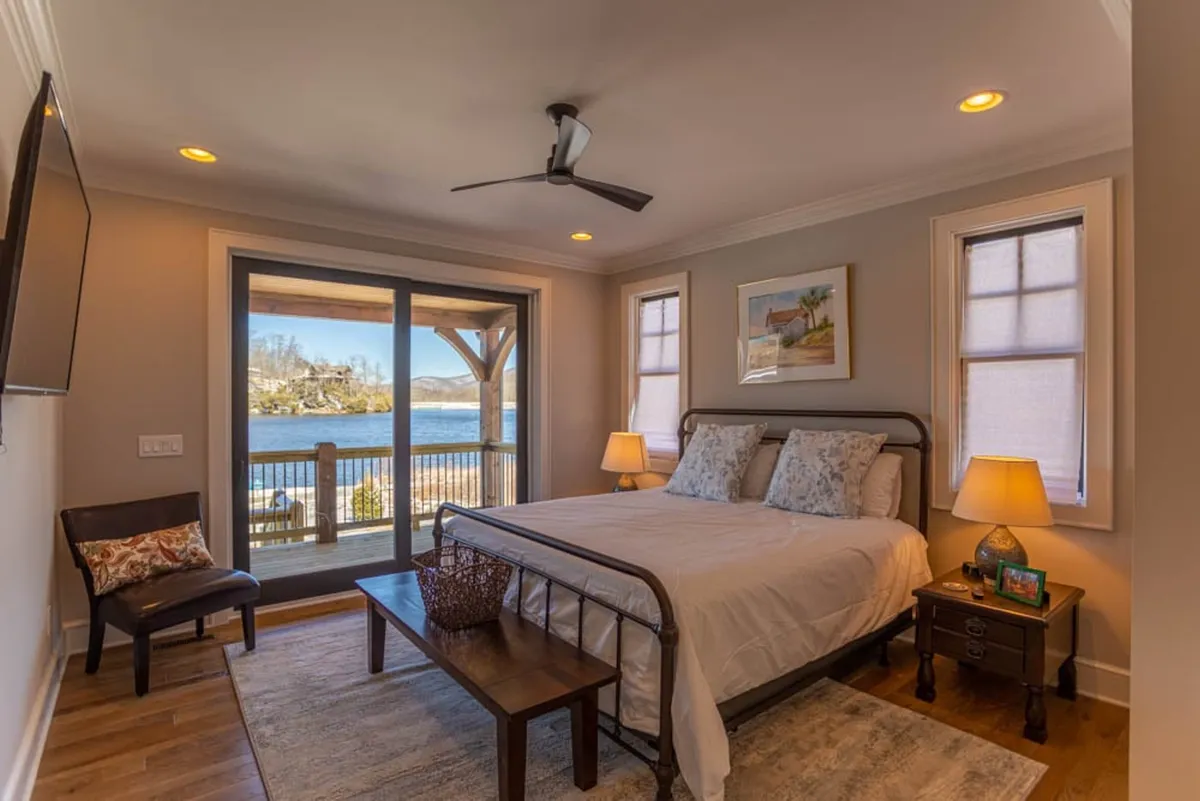 A serene bedroom with a plush bed positioned to face large windows that offer sweeping lake views. The room features a cozy nightstand, soft bedding, and a peaceful ambiance, emphasizing morning light and tranquility.
