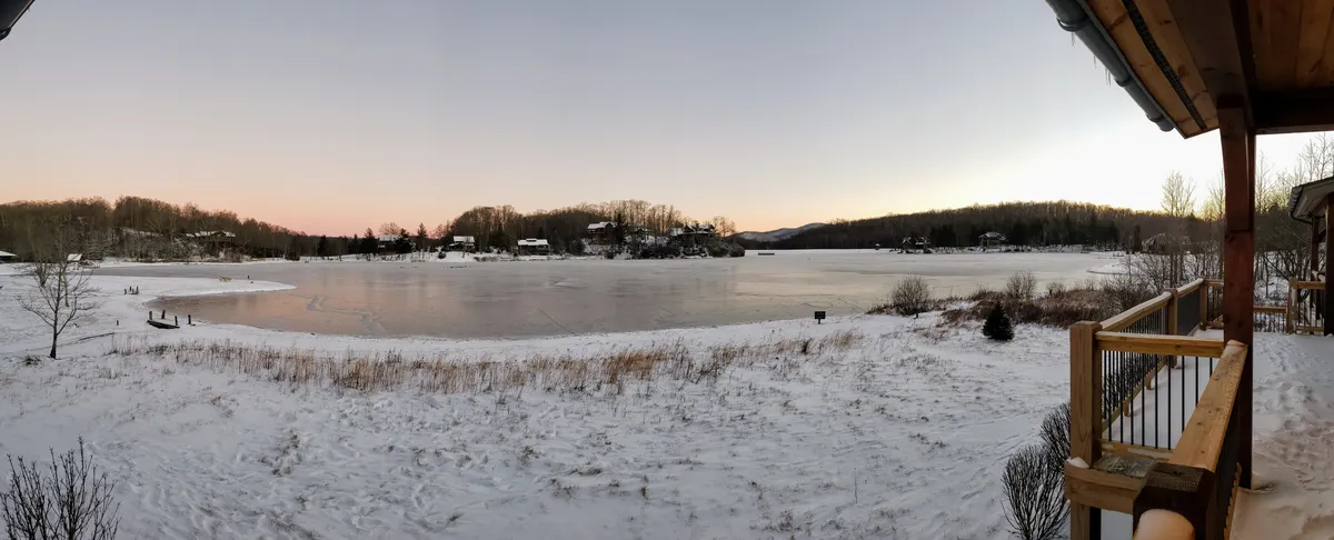 A winter landscape of a partially frozen lake, snow-laden trees, and cozy houses in the distance. The scene is tinted with the soft glow of sunset, evoking the magic of winter adventures and peaceful reflection by the water's edge.