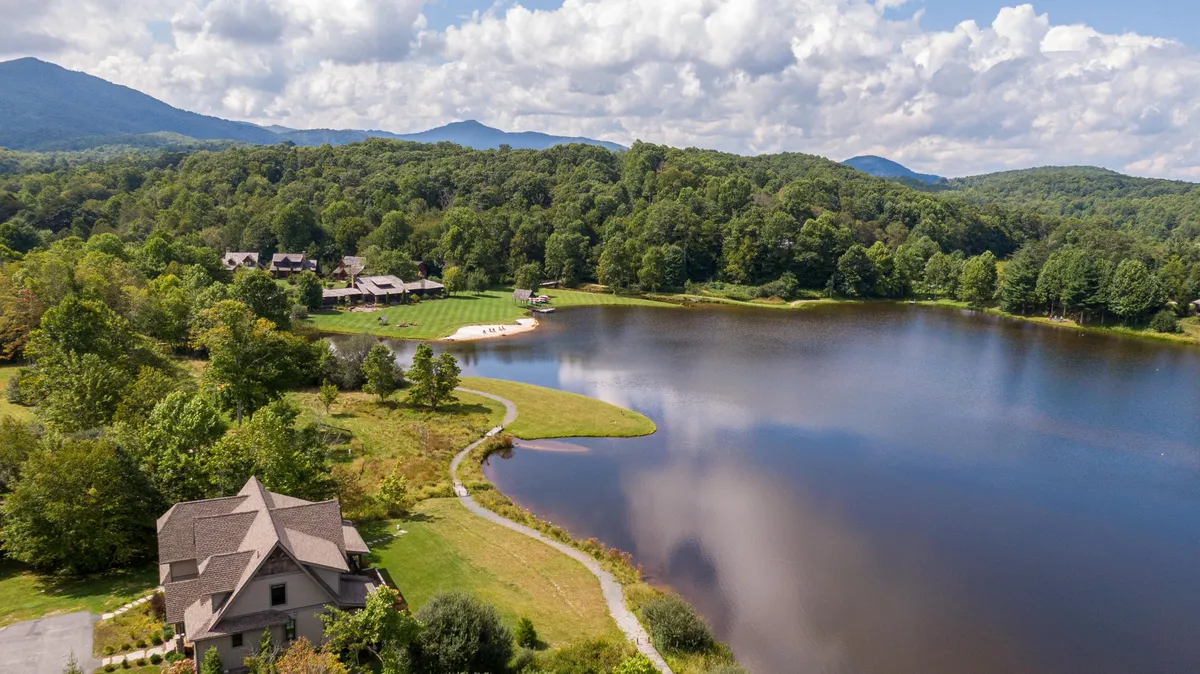 A wide, scenic view capturing the lake, surrounding mountains, lush greenery, and the home nestled in the landscape. The image offers perspective on the expanse of Sweetgrass, highlighting natural tranquility and the exclusivity of the location.
