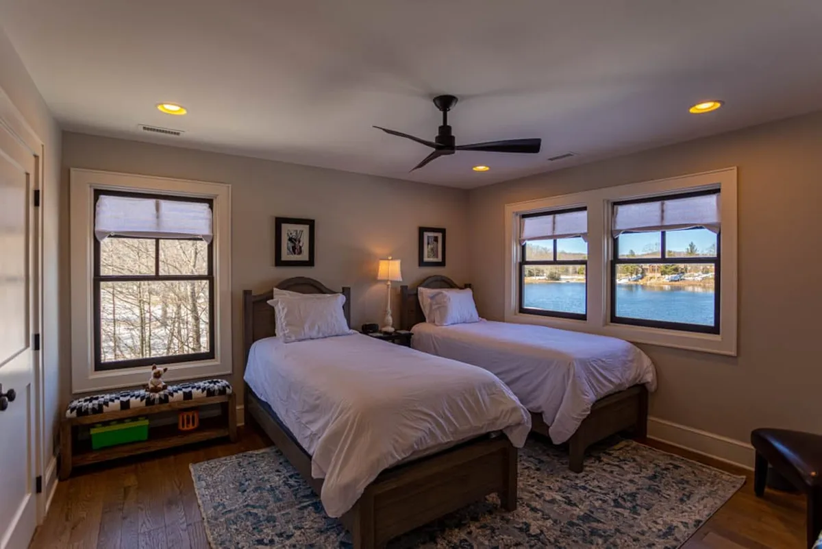 A cozy twin bedroom with two beds positioned by large windows overlooking water views. The room features decorative lamps and framed pictures, creating a welcoming space for families or friends to relax and enjoy the scenery.