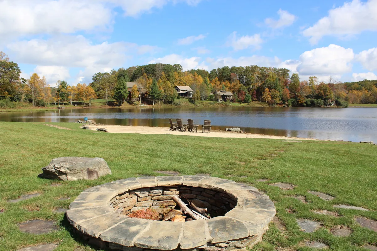 A cozy fire pit area with chairs set on a beach-like stretch beside the lake, surrounded by trees. The calm water and open sky create a welcoming space for campfire gatherings, swimming, and stargazing, with an emphasis on relaxation and connection to nature.