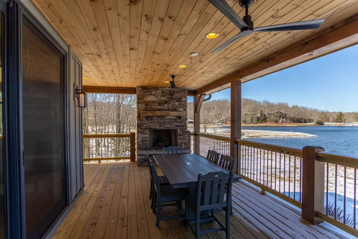 A cozy outdoor dining area on a spacious deck with a prominent stone fireplace and breathtaking views of the lake beyond. The dining table is set up for gatherings, while the fireplace invites warmth and relaxation during cooler evenings. The tranquil water and surrounding nature complete the scene, emphasizing all-season enjoyment.