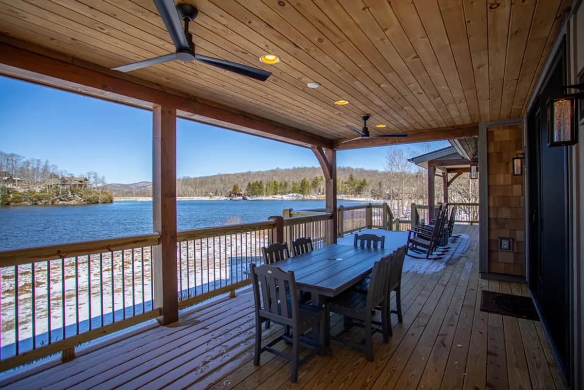 A serene lakeside porch with rocking chairs and a dining table, all oriented toward expansive lake views. The cozy wooden ceiling and tranquil setting suggest peaceful mornings with coffee, intimate meals, and stargazing sessions in the evening.