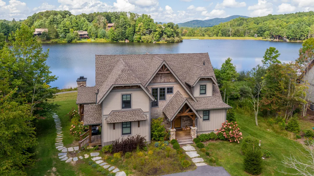 The image showcases a new-construction lakeside cabin nestled in the exclusive Sweetgrass community. The cabin sits just steps from the water’s edge, with manicured landscaping and a scenic pathway leading to the lake. The tranquil scene is enhanced by the warm glow of sunset reflected on the water, inviting outdoor relaxation and waterfront activities.