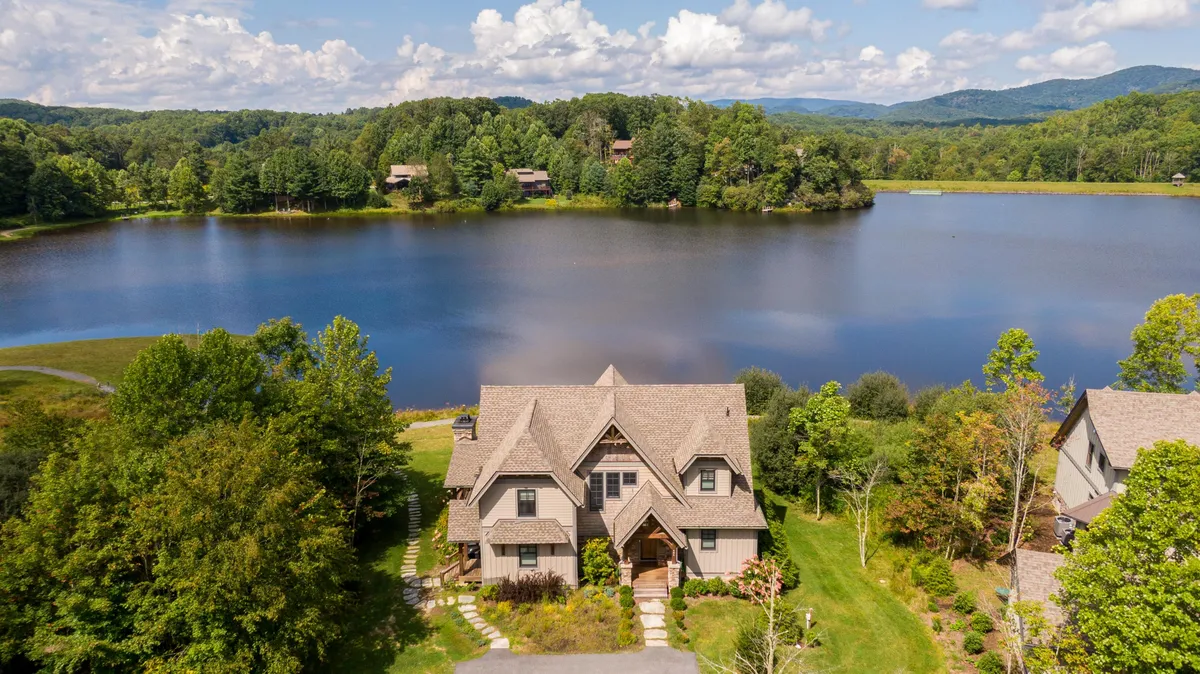 This exterior shot captures the house framed by trees and set beside a tranquil lake, with mountains visible in the background. The peaceful water and expansive views evoke a sense of escape and immersion in nature.