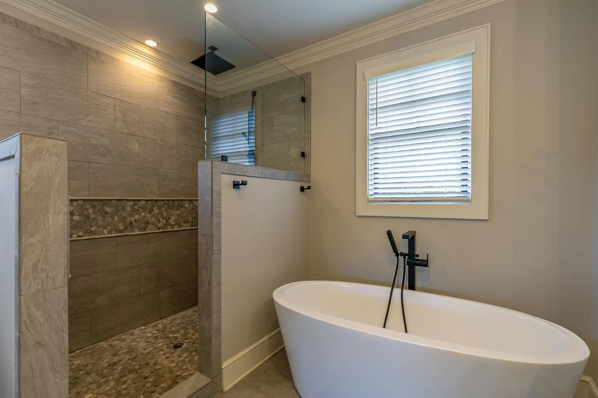 A luxurious bathroom featuring a freestanding soaking tub, a glass-enclosed custom tile shower, a textured stone accent wall, and soft, natural lighting filtering through a window. The atmosphere is calming and spa-like, inviting relaxation and self-care.