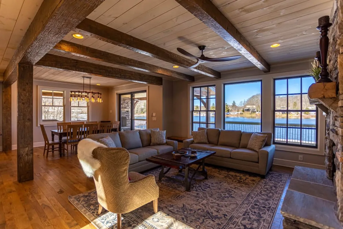This image features a spacious, open-concept living and dining area with a comfortable sofa, a dining table perfectly positioned to take in sweeping lake views through expansive windows, rustic exposed wooden beams overhead, and a striking stone fireplace. The scene is bathed in warm, natural light, creating an inviting and cozy atmosphere that encourages gatherings and relaxation.