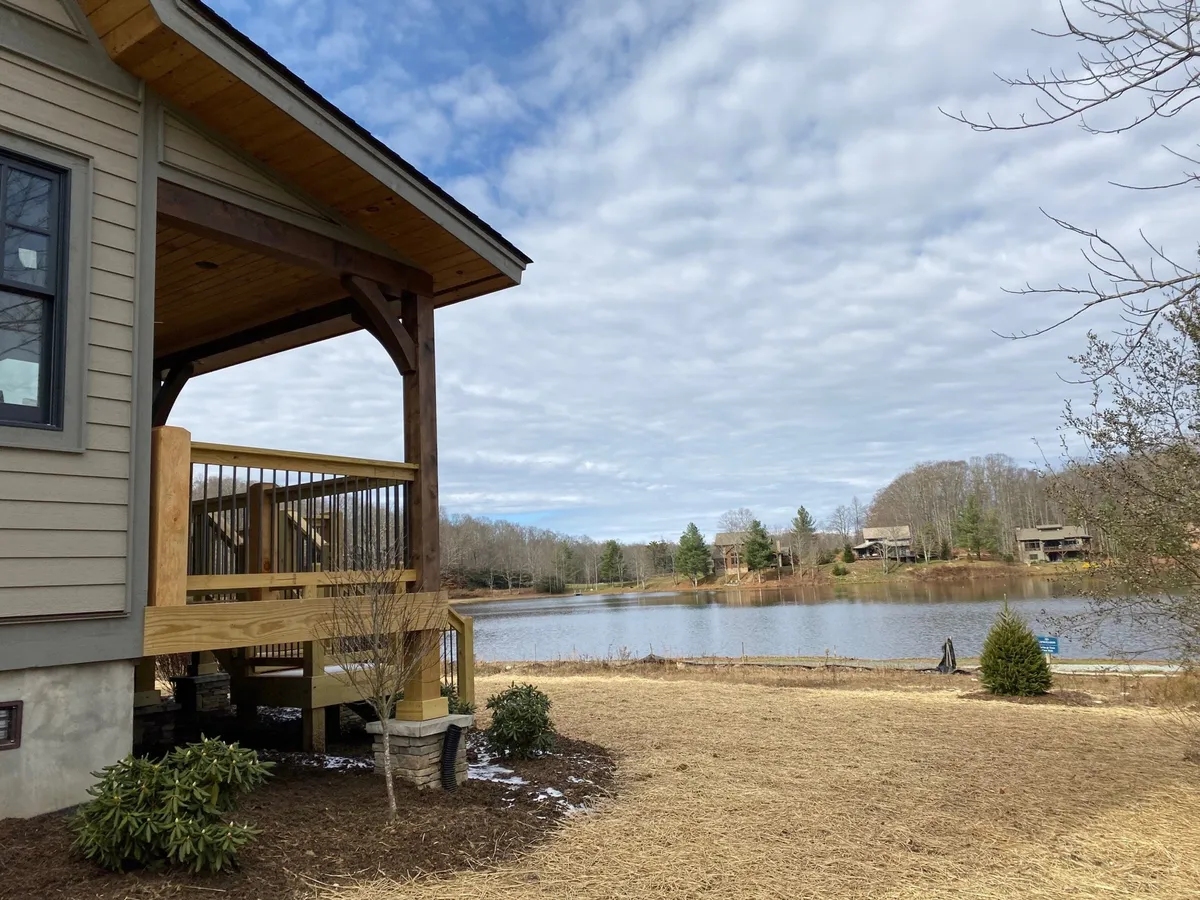 A serene lakeside scene featuring a spacious deck attached to the house, with a view of calm water, lush landscaping, and mountains in the distance. The deck is set up for relaxation and overlooks Sweetgrass's signature natural scenery, offering an inviting transition between indoor comfort and outdoor beauty.