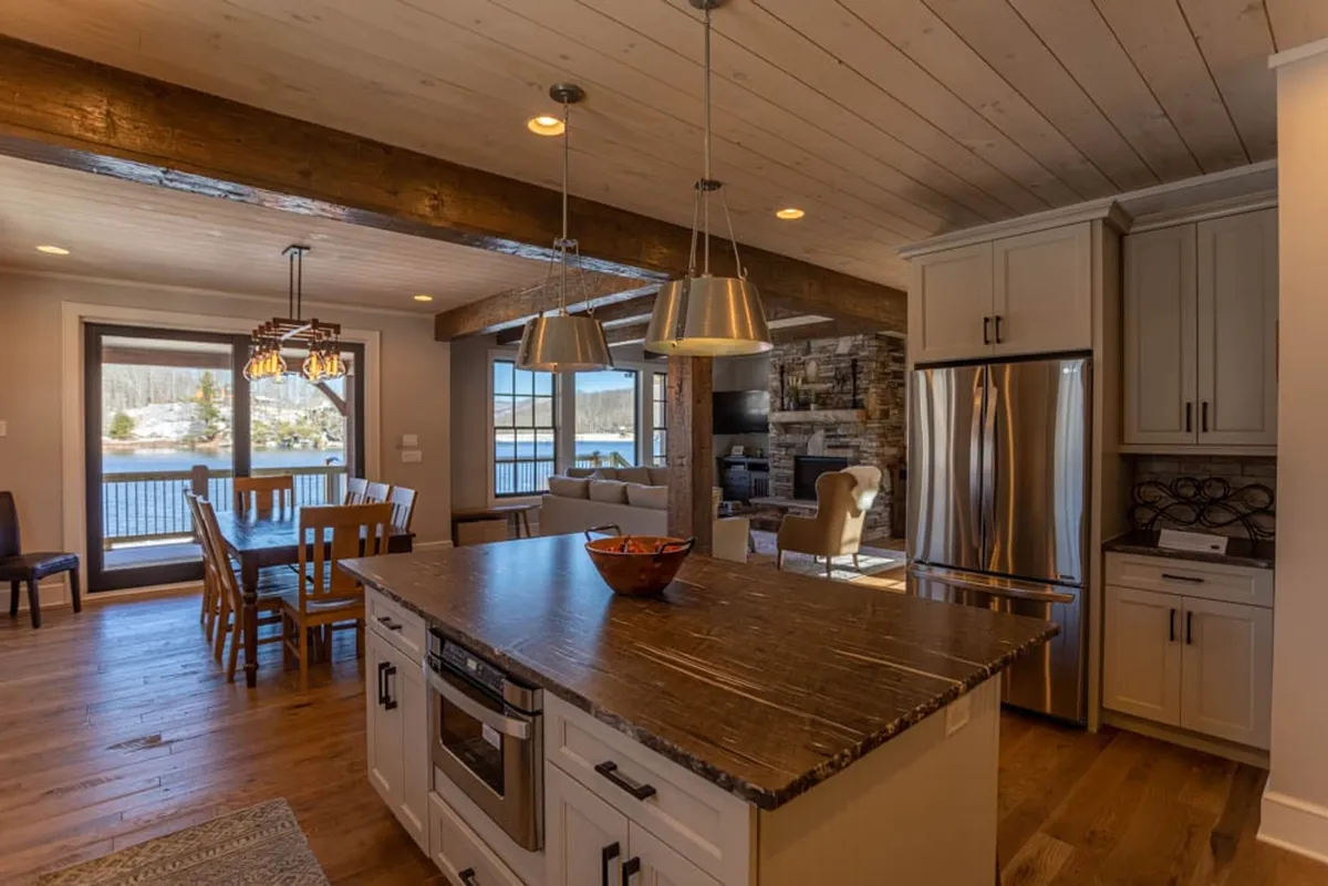 This image captures a seamless flow between the kitchen, dining, and living spaces. The kitchen island and dining table are both prominent, offering ample room for shared meals and activities. Large windows frame a serene water view, while a cozy fireplace anchors the room, reinforcing the sense of warmth and connection.