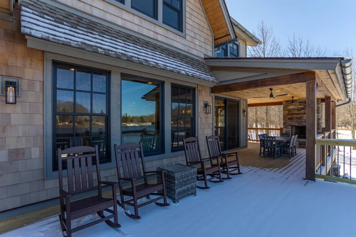 This cozy exterior view shows a wooden deck lined with classic rocking chairs and a prominent stone fireplace, set against a backdrop of large windows looking into the house. The deck offers ample space for outdoor dining, relaxing, and sharing stories, while the fireplace invites evening gatherings under a starry sky.