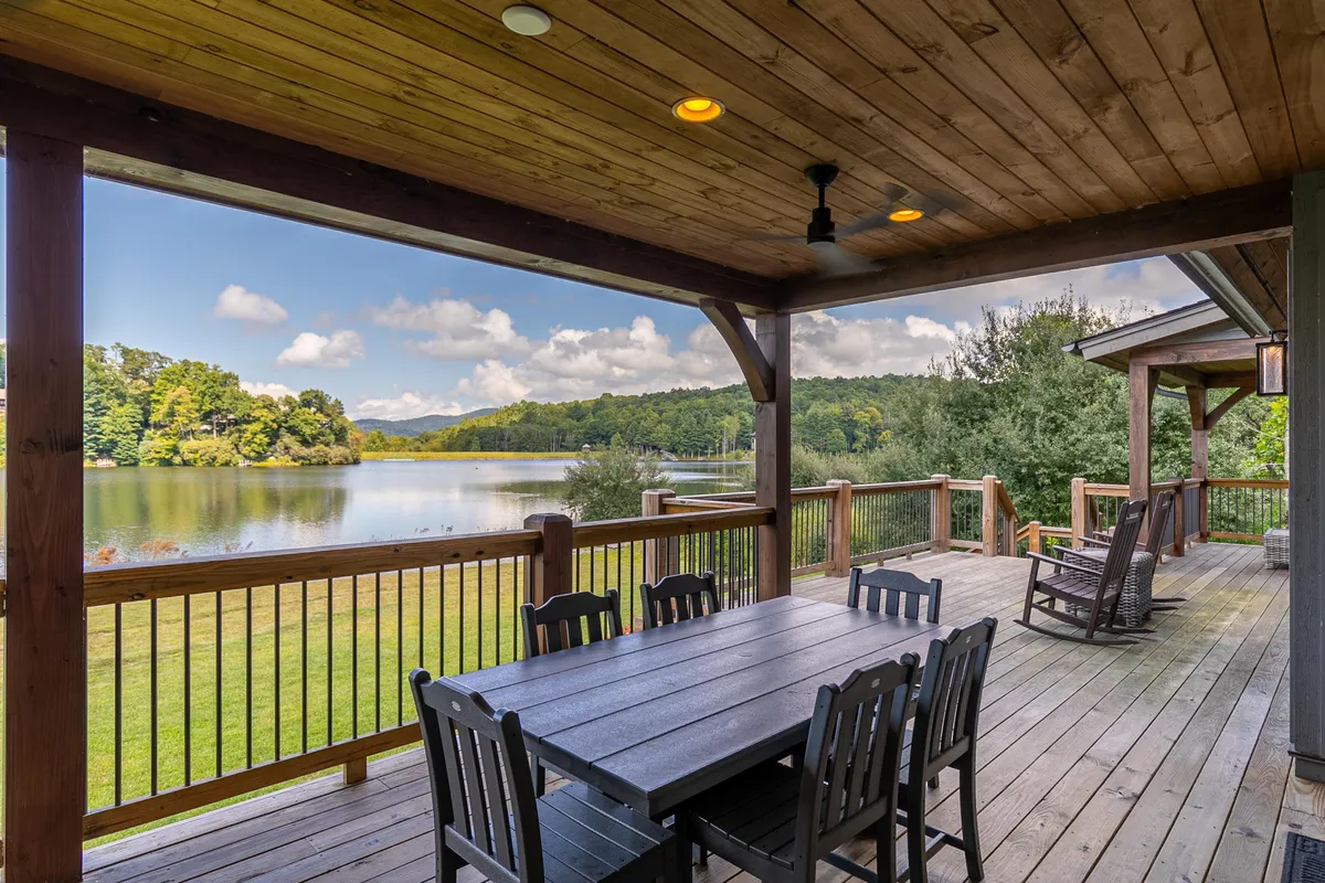 A spacious wooden deck overlooks a tranquil lake, with a dining table and rocking chairs inviting relaxation and group gatherings. The wooden railing frames the expansive water view, surrounded by lush greenery, suggesting a perfect spot for outdoor meals and socializing.