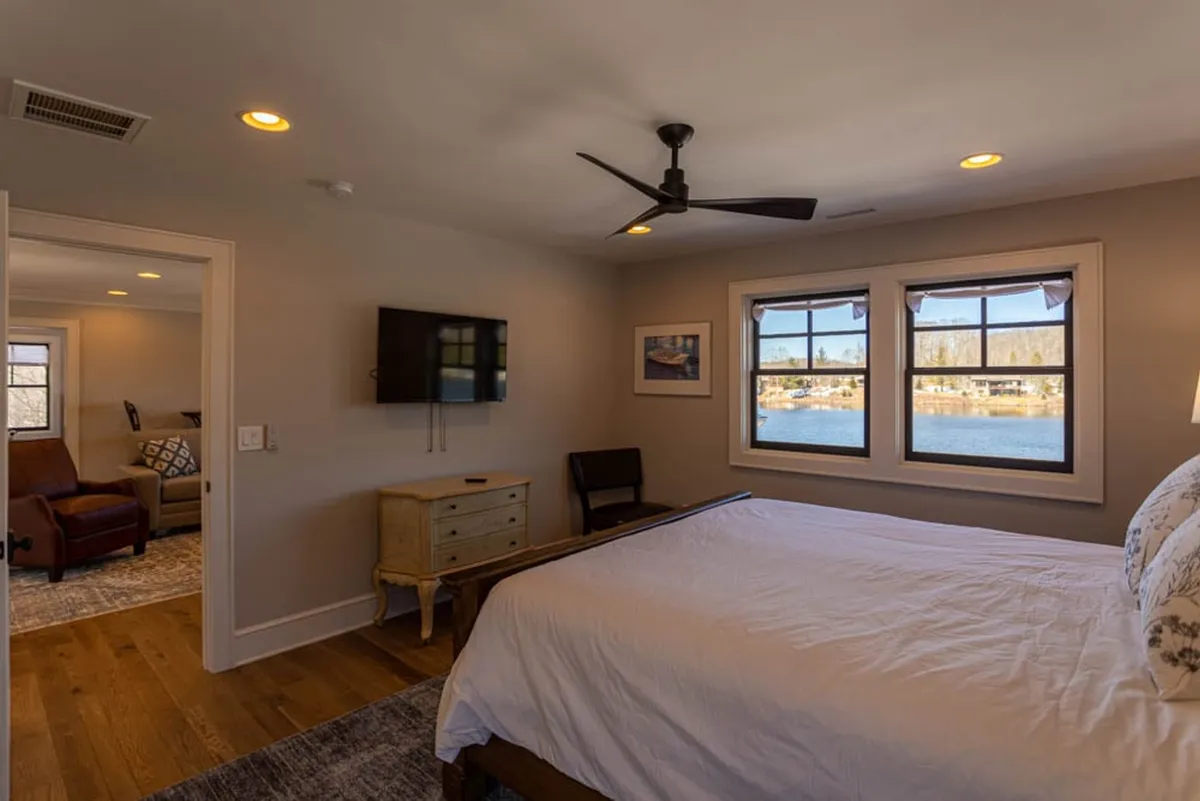 A tranquil bedroom furnished with a comfortable bed, an armchair near the window, decorative artwork, and a TV. The windows open to lake views, and the room radiates calmness and comfort, suggesting a restful experience.