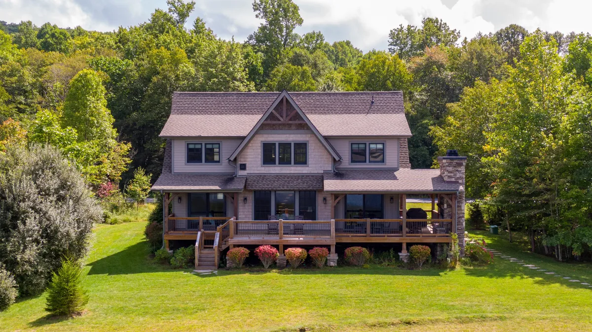 This image shows a wooden deck extending from the house, featuring large windows and a stone chimney, all set against lush greenery. The deck invites relaxation and outdoor gatherings, with the home’s architecture blending into its natural surroundings.
