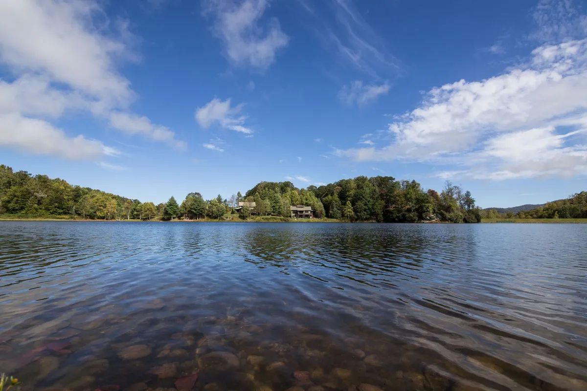 A calm lake surrounded by lush green trees, with a distant house nestled among nature. The scene is serene, evoking opportunities for fishing, boating, picnicking, and nature walks along the shore.