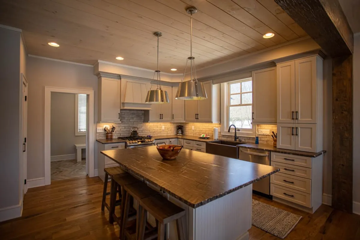 A warm, inviting kitchen scene featuring a large central island, modern hanging pendant lights, and gleaming stainless steel appliances. The island offers space for meal prep and casual dining, while the appliances and fixtures display the kitchen's high-end, functional style.
