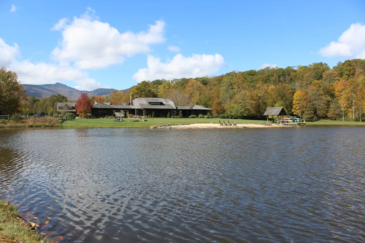 This image features the main house nestled by the lake, with a sandy area in the foreground and surrounded by mature trees. The tranquil water reflects the sky, while the setting highlights the peacefulness and natural beauty of the retreat, even as nearby development progresses.