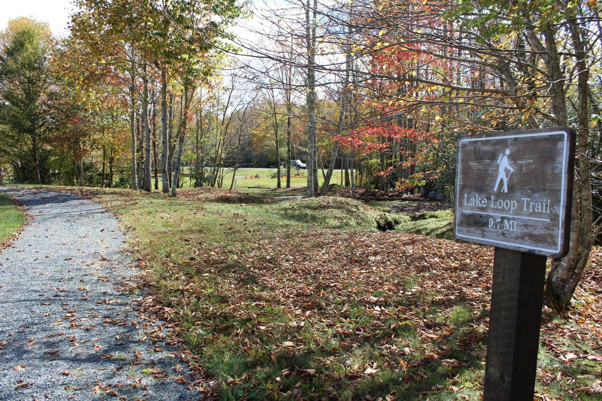 A tranquil exterior shot of the Lake Loop Trail sign marks the start of a scenic, winding path framed by mature trees. The path hints at further adventures and encourages families to set off on nature walks, birdwatch, and discover the beauty surrounding the lake.